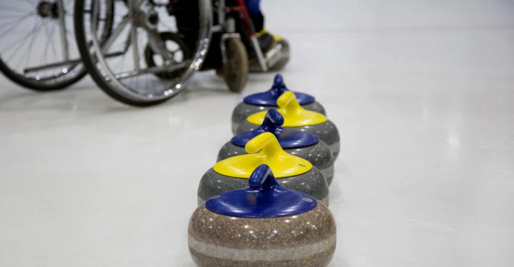 Wheelchair curler beside yellow-handled curling stones on the ice